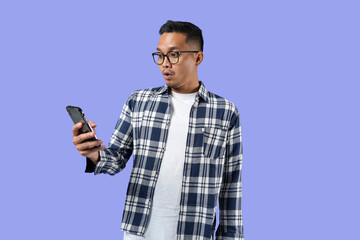Portrait of a young cheerful excited Asian man standing isolated over a purple background, using a mobile phone