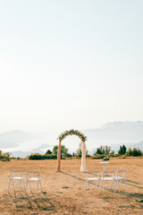 White chairs stand in front of a wedding arch and a reception table with a bouquet of flowers in a...
