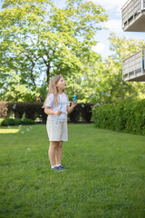 8 year old blonde girl blowing soap bubbles on the street, on a warm sunny day, full length photo in light clothes, a lot of greenery in the background