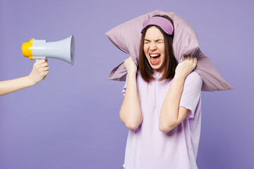 Young sad mad woman wear pyjamas jam sleep eye mask at home hold megaphone cover ears with pillow...