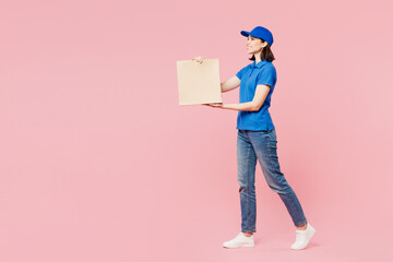Full body side view delivery girl employee woman wear blue cap t-shirt uniform workwear work as dealer courier hold brown blank craft paper bag go isolated on plain pink background. Service concept.