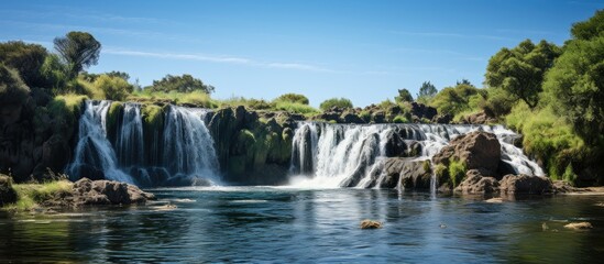 Naklejka premium A stunning waterfall featured in a scenic landscape with a peaceful surrounding and a clear blue sky in the background is captured in a copy space image