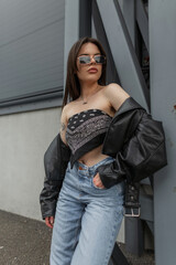 Cool fashion beautiful girl woman in fashionable clothes with bandana top with leather jacket and jeans posing on the street near metal construction