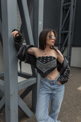 Fashionable beautiful urban hipster girl with sunglasses in fashion casual clothes with a bandana top, leather jacket and jeans posing in the city near a metal black building