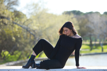 Young and beautiful woman, brunette and dressed in black sitting on a wall in the park, in the...