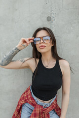 Cool stylish young girl with a tattoo in fashion clothes with a T-shirt, shirt and jeans adjusts her glasses and stands on a concrete background