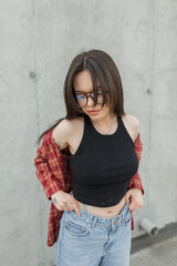 Young fashionable girl student with glasses in fashionable denim clothes with a shirt on the street near a concrete wall