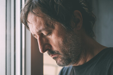 Downhearted man with eyes closed leaning his head close to the apartment window. Depressed unhappy male sad and alone at home.