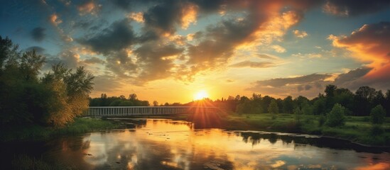 A picturesque sunset with the sun setting over the horizon casting golden rays on a bridge over a river highlighting a lush green clearing with tall trees under a sky painted with colorful clouds Copy - Powered by Adobe
