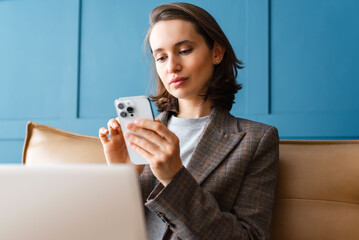 Beautiful woman with brown hair sitting on the sofa, using on mobile phone and working on laptop.