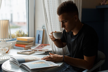 young man sits in a cafe near the window, drinks coffee and reads a book. Rest and reading