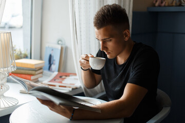 Handsome young man sits in a coffee shop by the window, drinks coffee and reads a book