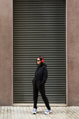 Portrait of a young woman in a black hoodie, red headphones, and sunglasses, listening music on a cool, urban street style. 