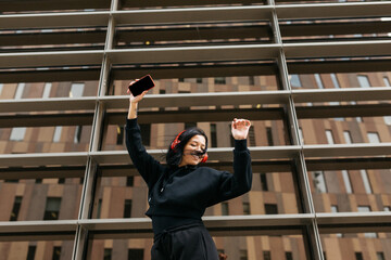 Young woman in a black hoodie dancing with headphones on and a phone in her hand, against the...