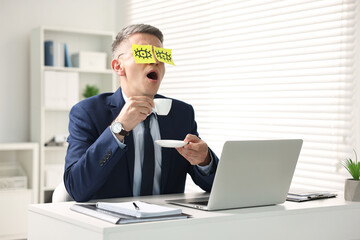 Man with fake eyes painted on sticky notes holding cup of drink and yawning at workplace in office