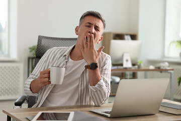 Man with cup of drink yawning at wooden table in office