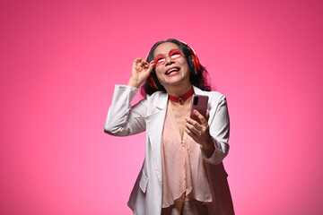 Laughing senior woman in headphone with mobile phone standing against pink background
