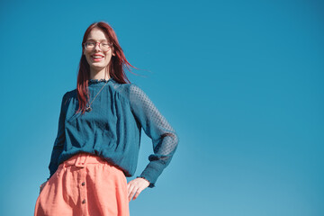 Smiling teenage girl with red hair and glasses wearing blue blouse and coral skirt. Outdoor portrait with clear blue sky background. Close-up shot with copy space.