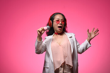 Stylish elderly woman in glasses and headphones singing with microphone over pink background
