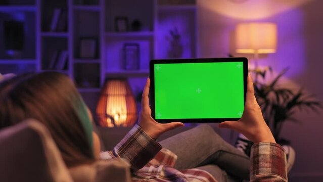 Closeup shot of girl lying on sofa using tablet in horizontal position with green screen mockup in evening room using different gestures while watching video.