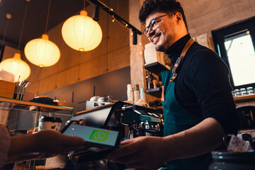 Young woman paying to the bartender for a takeaway coffee with the smartphonephone.