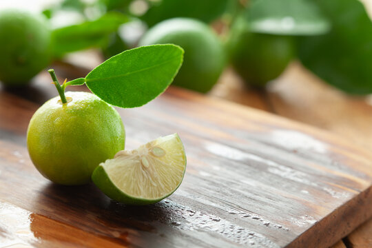 Close-up of fresh green organic whole and sliced lemons (Citrus limon) with leaves, on a wooden chopping board.
