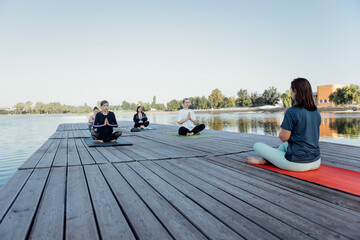 Outdoor yoga class with an instructor near the river. Young women sit cross-legged on mats