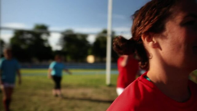 Female Rugby Player Holding Ball on the Field - A female rugby player in a red jersey holding a rugby ball, with teammates in the background on the field.
