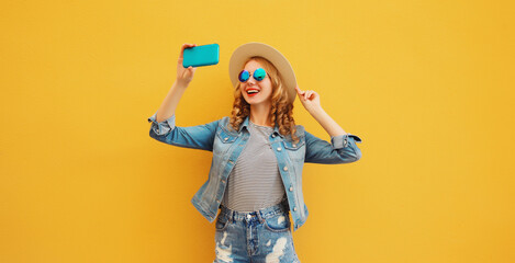 Happy smiling young woman taking selfie with smartphone wearing a summer hat on yellow background