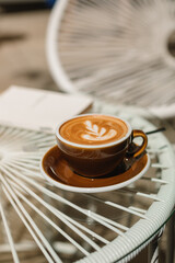 A cup of cappuccino and a book on a glass table on the terrace on a sunny summer morning.