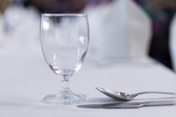 Close-up of a clear wine glass with a traditional round shape. On a white dining table with eating utensils, there is a blurry background of the restaurant