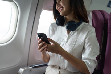 Beautiful Asian in aeroplane. working, travel, business concept Traveler on Plane Using Smartphone with Headphones, Smiling and Happy