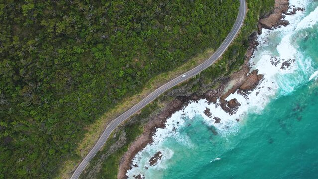 4K Drone ariel video of a camper van driving along the Great Ocean Road. View of green nature on one side of the road and the blue ocean and waves crashing on the other side of the road.
