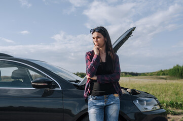 A young sad beautiful woman stands near a broken down black car after an accident with the hood...