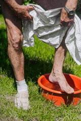 Adult elderly man gardener washes his feet in a red basin, a bowl of water on the green grass after...