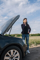 A young beautiful lonely woman, a brunette driver, stands near a broken down black car with an open...