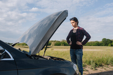 A young sad beautiful woman stands near a broken down black car after an accident with the hood...