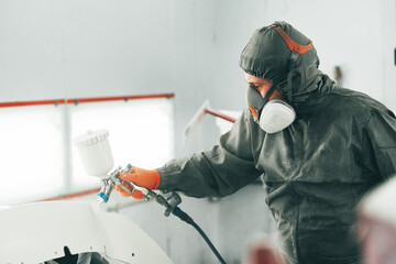 Male painter in respiratory mask and protective suit painting car with spray gun in car service