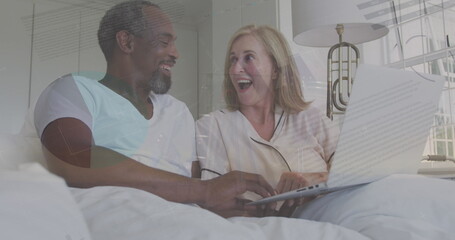 Image of statistical data processing over happy biracial couple using laptop in the bed at home