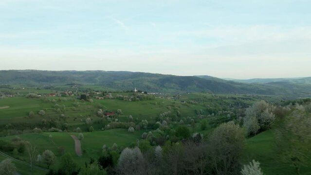 Drone glides low over blooming trees in Central Slovakia's Hrinova area, revealing a modern Catholic church atop a hill. LuPa Creative.