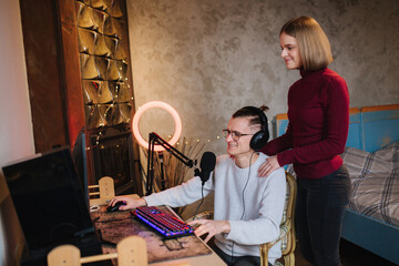 Young attractive wife help husband to work. Handsome man sits at the table and work on computer. Man using professional microphone and headphones