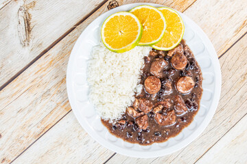 A delicious plate of Brazilian feijoada with white rice and orange slices served on a white plate against a rustic wooden background.