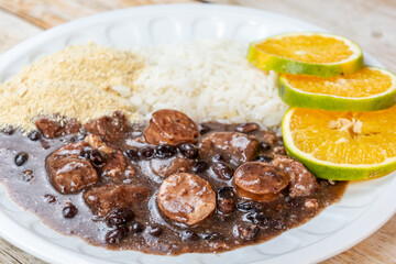 Traditional Brazilian feijoada with black beans, sausage, rice, farofa, and orange slices on a white plate, ready to be enjoyed.