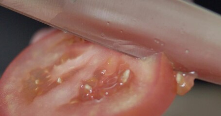 Super-closeup footage of adult hands cutting a tomato slice with sharp knife on a cutting board