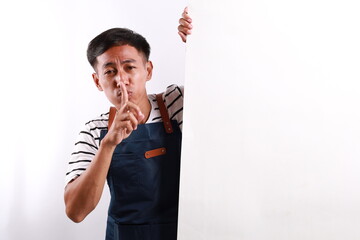 Asian man in blue apron peeking behind wall with silent gesture