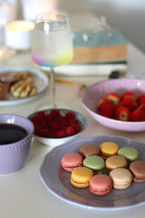 Plate of pastel macarons, cookies and chocolate, cup of tea of coffee, glass of bubble water, various berries, books and accessories on the table. Selective focus, pastel colors.