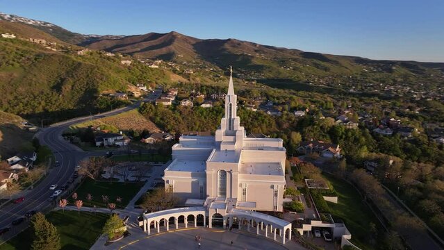 Drone orbiting shot of Orem Utah Temple at sunset, with a mountain road and green landscape in Utah