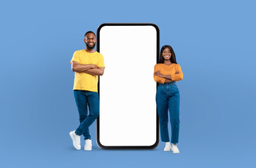 Black couple positioned next to a blank screen while standing against a blue background. They...