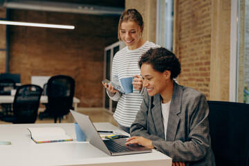 Two smiling female coworkers working on project together and use laptop sitting in office