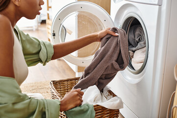 African American woman with afro braids putting a cloth into a dryer in a bathroom during laundry...